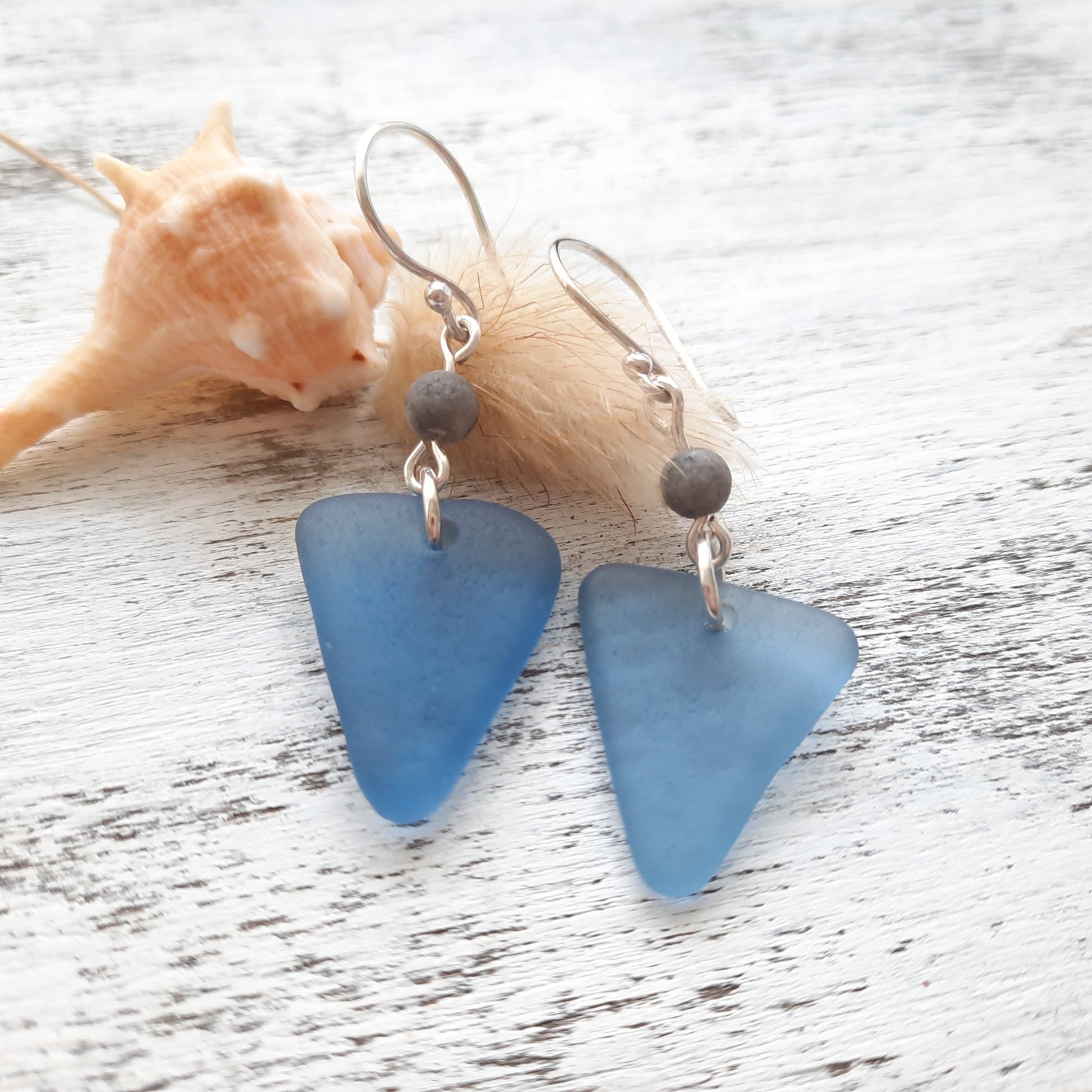 Blue sea glass earrings with silver hooks on a wooden surface with a shell.