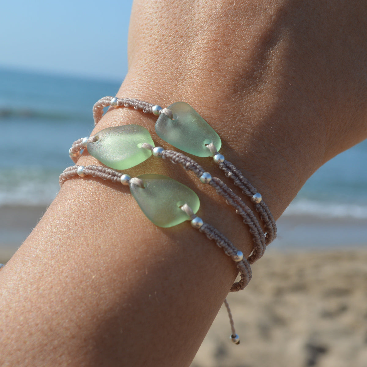 A close-up of a person's wrist wearing three layered bracelets withsea foam green sea glass pendants and beige cord, photographed outdoors on a sandy beach with the ocean in the background.