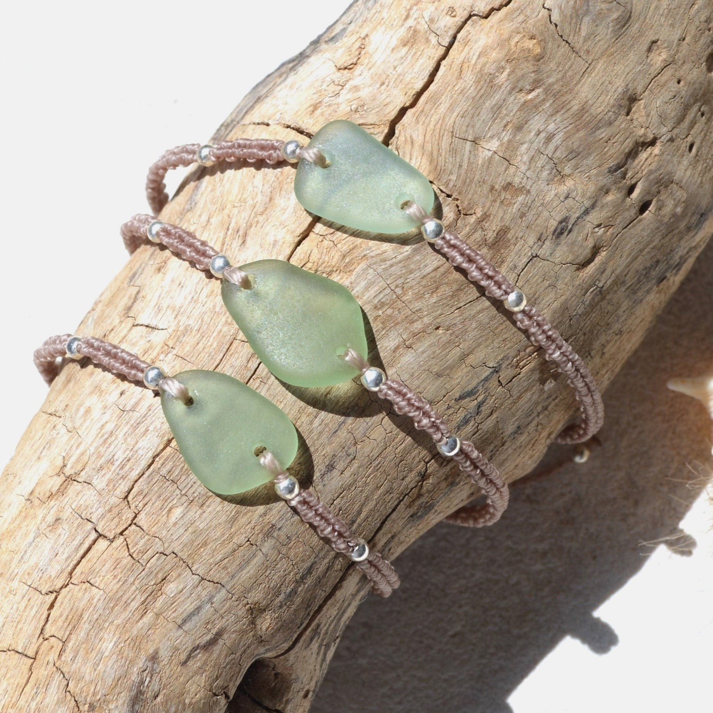Three sea glass beaded woven bracelets with seafom green sea glass and beige cord, displayed on a piece of driftwood.